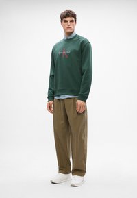 Sweatshirt verde com logotipo, sobre uma camisa listrada azul, combinado com calças largas bege e tênis brancos. Design minimalista.