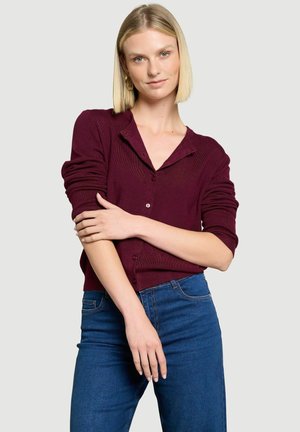 Žena s krátkymi blond vlasmi, oblečená v bordový zapínací sveter a modré džínsy, stojaca s prekríženými rukami na svetlom pozadí.