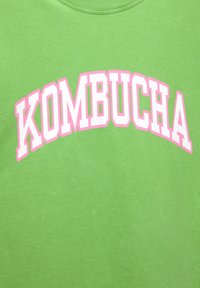 T-shirt in cotone verde con testo audace e arcuato che legge "KOMBUCHA" in bianco con contorno rosa. Texture liscia, design a collo rotondo.