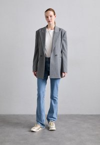 Blazer grigio oversize con revers a scollo a italia, doppi bottoni e tasche laterali, indossato su una t-shirt bianca e jeans di denim chiaro. Sneakers con dettagli metallici.
