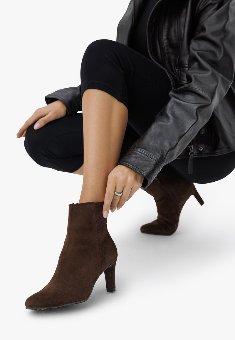 Rjave nobučne gležnjarje z ostrim nosom in 4-palčnim peto, ki imajo zadrgo in gladko teksturo, postavljene na belo ozadje.