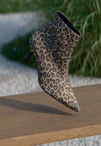 Stivale alla caviglia a stampa leopardata con punta affusolata e tacco sottile appoggiato su una superficie di legno, con piante verdi e ghiaia sullo sfondo.
