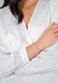 Silberarmband mit abwechselnden blauen Edelsteinen, am Handgelenk getragen, kombiniert mit einer weißen Bluse mit langen Ärmeln und gerafften Manschetten.
