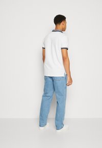 Cămașă polo albă cu guler tăiat în navy, purtată cu blugi din denim de culoare bleu deschis și adidași albi. Croi lejer, material moale și design simplu.