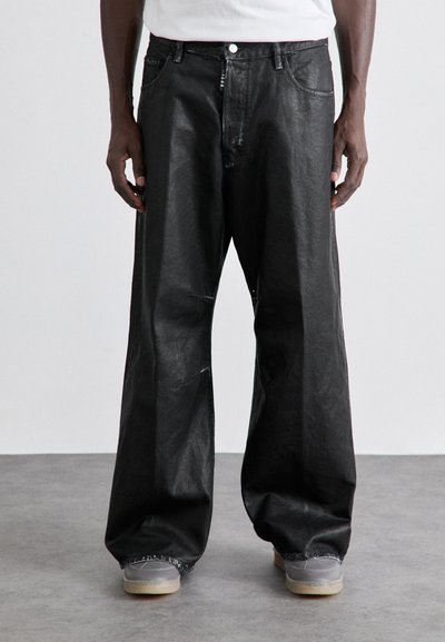 Pantalons en denim noirs à jambe large avec une texture lisse, design à cinq poches et détails de couture blanche, portés avec des chaussures grises sur un fond uni.