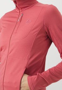 Primer plano de una persona con chaqueta atlética roja con cremallera, con un bolsillo en el pecho con cremallera y la mano en el bolsillo lateral, sobre fondo blanco.