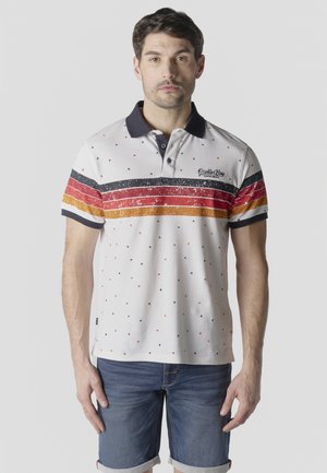 Hombre con camiseta tipo polo blanca con rayas horizontales en negro, rojo y naranja y pequeños puntos de colores, combinada con pantalones cortos de mezclilla enrollados.