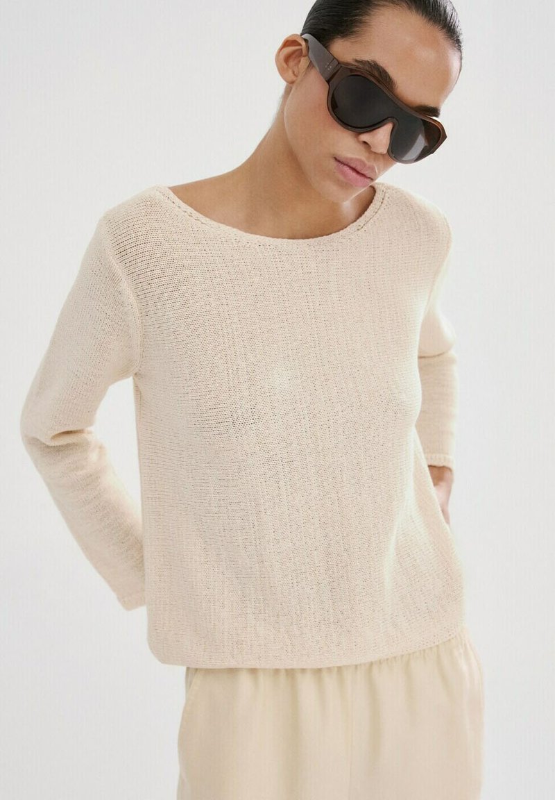 Femme portant de grosses lunettes de soleil foncées et un pull en maille crème ample avec un pantalon beige clair sur un fond blanc.
