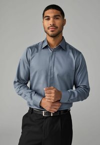 Camisa de manga larga en azul claro, de textura suave, combinada con pantalones negros y una hebilla metálica, manos entrelazadas a la altura de la cintura.