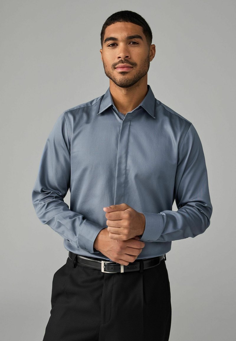 Camisa de manga larga en azul claro, de textura suave, combinada con pantalones negros y una hebilla metálica, manos entrelazadas a la altura de la cintura.