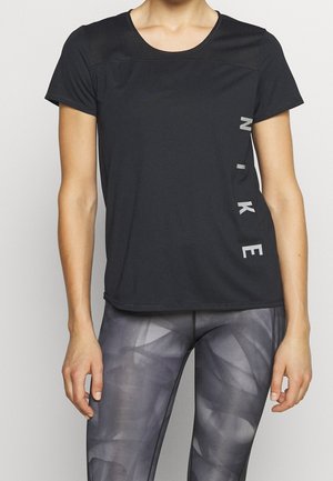 Vrouw draagt een zwart Nike T-shirt met korte mouwen en een verticaal logo aan de zijkant en grijze patroonleggings, staand met ontspannen armen.