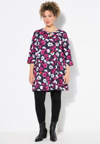 Bloemen tuniekjurk in marineblauw, met roze en witte bloemen, een keyhole-halslijn, driekwart mouwen, gecombineerd met zwarte legging en enkellaarsjes.