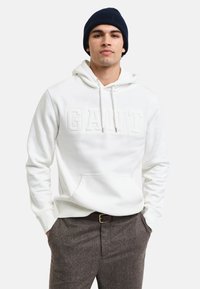 Sudadera blanca con el logo "GANT" en relieve, cordones y bolsillo tipo canguro, combinada con pantalones marrón texturizados y un gorro azul marino.