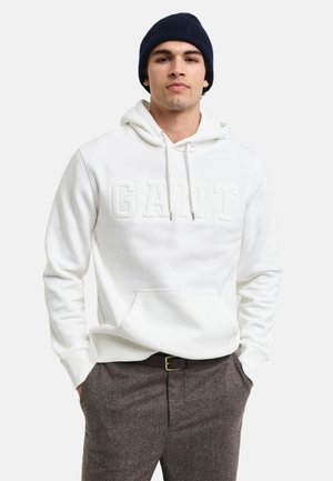 Sudadera blanca con el logo "GANT" en relieve, cordones y bolsillo tipo canguro, combinada con pantalones marrón texturizados y un gorro azul marino.