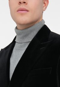 Primo piano di una persona che indossa un maglione grigio a collo alto sotto un blazer di velluto nero, mostrando la parte inferiore del viso e la zona del collo.