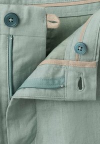 Gros plan de la ceinture d’un pantalon vert clair montrant un bouton, une boutonnière, une fermeture éclair et des détails de couture beige.