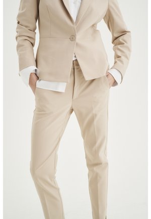 Beige tailored suit with single-button blazer and matching slim-fit trousers, worn with a white shirt, hands in pockets.