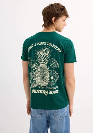 Camiseta de algodón verde con un gran gráfico de piña blanca y el texto "Entrega de Frutas y Jugos" y "Jugo de Piña Fresca y Rica" en la parte trasera.
