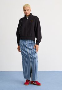 Zwarte cropped sweatshirt met een kwart rits, gecombineerd met een lange rok in het blauw met verticale strepen, met zijsplitten en zakken. Rode sneakers.