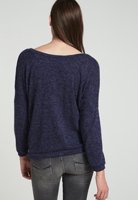 Marinblå, texturerad tröja med vid halsringning, sänkt axelsöm och långa ärmar, kombinerad med grå, åtsittande jeans.