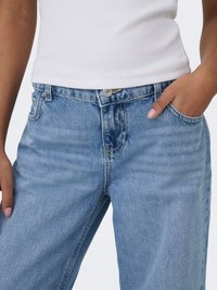 Bela majica brez rokavov, nošena z visokim pasom svetlo modrih kavbojk iz jeansa, ena roka v žepu, obrezano od ramen do stegnenic.