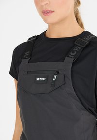 Schwarze isolierte Latzhose mit verstellbaren Trägern, einer mit Reißverschluss versehenen Brusttasche und einem strukturierten Stoff, der auf der Vorderseite ein Logo-Patch trägt.