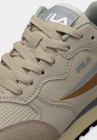 Gros plan sur une chaussure de sport Fila beige montrant le tissu texturé, les surpiqûres en cuir, les lacets beige et le logo sur le côté et la languette.