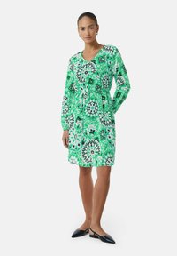 Groene jurk met patroon, lange mouwen, V-hals en een gecinchde taille. Bevat bloem- en geometrische ontwerpen met zwart-witte accenten.