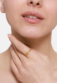 Bague en or avec un design de croix texturé, portée à l'index. Une peau lisse et une couleur de lèvres naturelle sont visibles en gros plan.