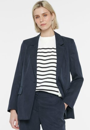 Frau mit kurzen blonden Haaren, die einen marineblauen Blazer und passende Hosen über einem weißen Hemd mit schwarzen Horizontalstreifen trägt.