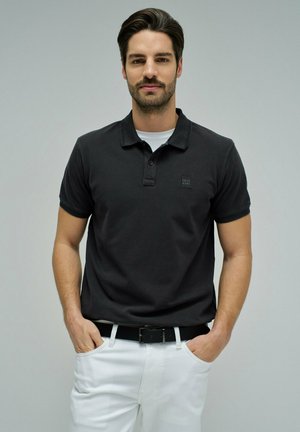 Homme aux cheveux noirs et barbe portant un polo noir sur un t-shirt blanc, un pantalon blanc et une ceinture noire, les mains dans les poches.