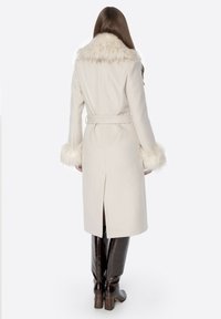 Długi kremowy płaszcz z kołnierzem i mankietami z sztucznego futra, z paskiem w talii, rozcięciem z tyłu, noszony na brązowych kozakach do kolan. Gładka faktura.