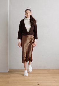 Cardigan fuzzy maro, pulover alb cu guler, fustă satin maro și adidași albi. Modelul stă pe un podea din lemn, în fața unui perete gri deschis.