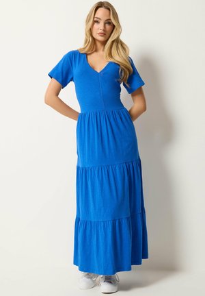 Femme aux longs cheveux blonds portant une robe maxi bleue à volants et des baskets blanches, debout devant un fond clair uni.