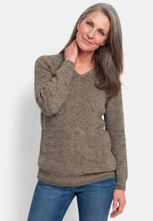 Frau mit langen grauen Haaren, die einen braunen Strickpullover und blaue Jeans trägt, lächelt sanft mit einer Hand am Hals.