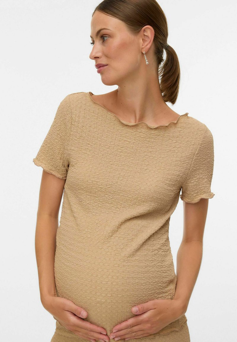 Donna incinta in un top beige a maniche corte con una texture, mentre tiene delicatamente la sua pancia e guarda di lato su uno sfondo semplice.
