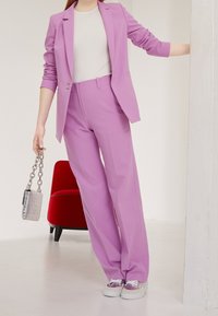 Traje ajustado de color púrpura claro con mangas largas y pantalones de pierna ancha, combinado con una blusa blanca y zapatillas con detalles plateados. Sosteniendo un bolso plateado.