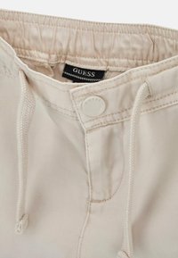 Pantaloni beige con vita regolabile tramite cordoncino, chiusura a bottone e etichetta con marchio. Il tessuto presenta una texture liscia con dettagli di cucitura fine.