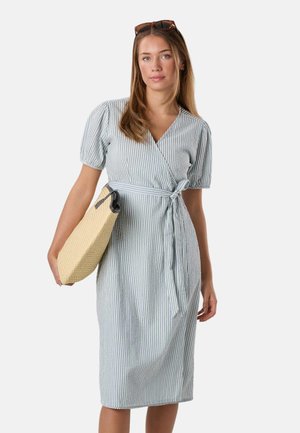 Kvinde iført en lyseblå og hvid stribet slå-om-kjole, der holder en beige vævet clutch taske, med solbriller på hovedet, mod en ensfarvet baggrund.