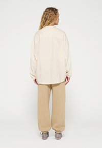 Overmaat crèmekleurig shirt met een knoopsluiting op de kraag, gecombineerd met geribbelde beige wijde broek en grijze sneakers. Zachte textuur, relaxte fit.