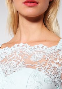 Robe bleu clair avec une superposition de dentelle ornée de motifs floraux. L'encolure est festonnée, mettant en valeur un brodé compliqué et des textures délicates.