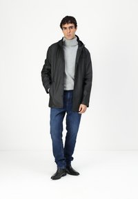 Veste noire imperméable avec capuchon, fermeture à boutons et poches latérales, portée sur un col roulé gris et un jean bleu, associée à des bottes noires.