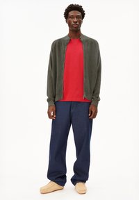 Grüne Zip-Jacke, rotes T-Shirt, lockere blaue Jeans und beige Schuhe, getragen von einem Modell mit kurzen lockigen Haaren vor einem weißen Hintergrund.