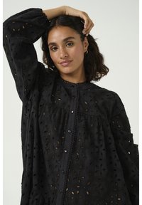 Svart broderad klänning med en texturerad, öppen mönstring, långa ärmar, knäppning fram och en rundad krage med dekorativt kant.