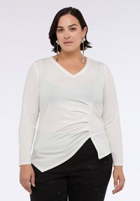 Top bianco a maniche lunghe con scollo a V, dettagli drappeggiati ai lati e accenti scintillanti sottili. Abbinato a pantaloni neri con texture.
