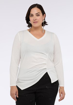 Blusa blanca de manga larga con escote en V, detalle drapeado en los lados y sutiles toques brillantes. Combinada con pantalones negros texturizados.