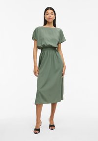 Vestido midi verde con mangas cortas, cintura elástica fruncida y textura suave. Llevado con tacones negros de punta abierta.