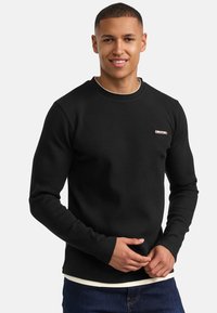 Zwarte geribbelde langemouwen shirt met een ronde hals en een klein logo op de linkerkant van de borst, gecombineerd met een witte onderhemd en denim spijkerbroek.