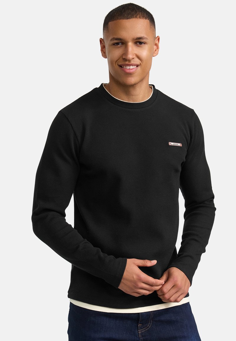 Zwarte geribbelde langemouwen shirt met een ronde hals en een klein logo op de linkerkant van de borst, gecombineerd met een witte onderhemd en denim spijkerbroek.