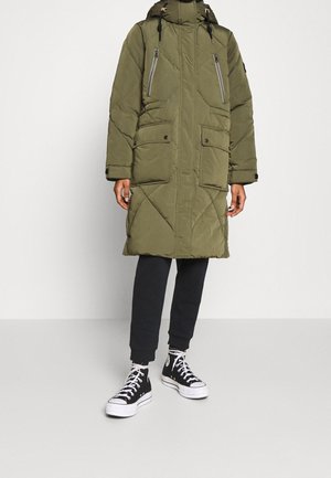 Vue de face d'une personne portant un manteau matelassé vert olive longueur genoux avec capuche, un pantalon de jogging noir et des baskets montantes noires avec des lacets blancs.